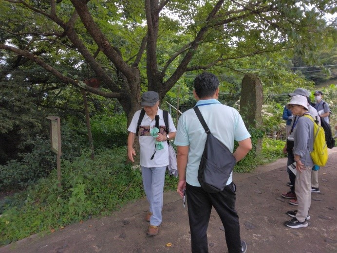 草木の生い茂る道路脇に置かれた念仏橋の板碑を見る受講者たちの写真