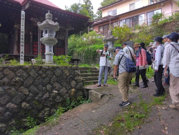 羽黒観音の坂の下を差しながら説明している講師と話を聞く受講者たちの写真
