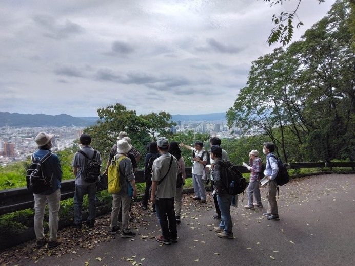 曇り空と綺麗な街並みを見ながら福島盆地について説明する講師と聞く受講者たちを後ろから撮影した写真
