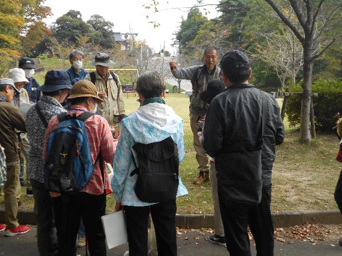 公園内にある欅の葉について説明する講師と話を聞く受講者たちを撮影した写真