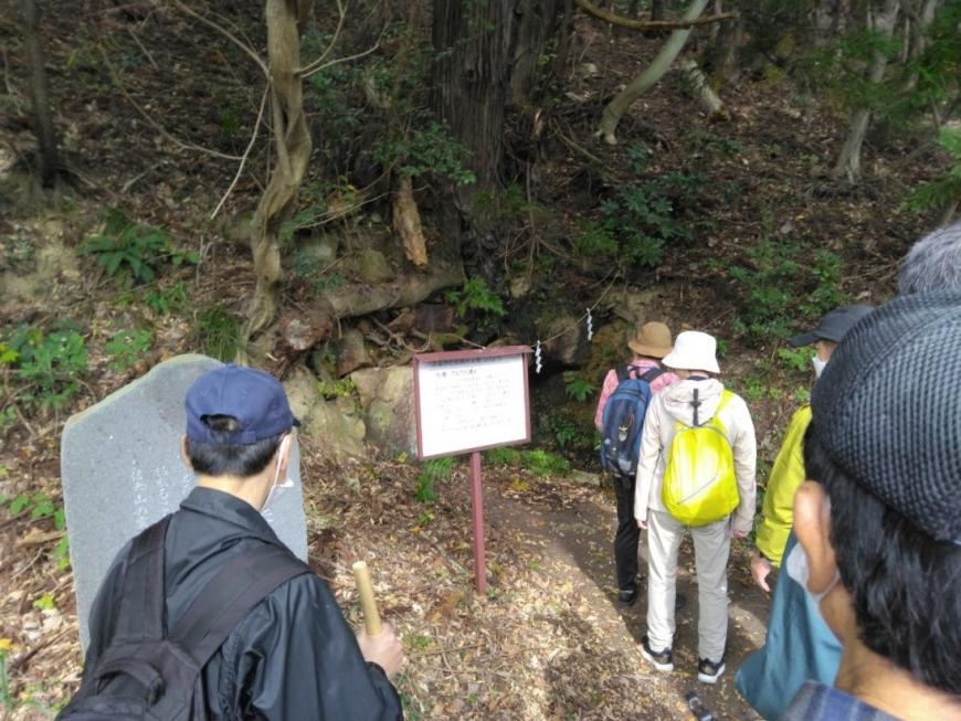 森の開けた場所に置かれた、たんたら清水の看板を読んでいる受講者たちを撮影した写真