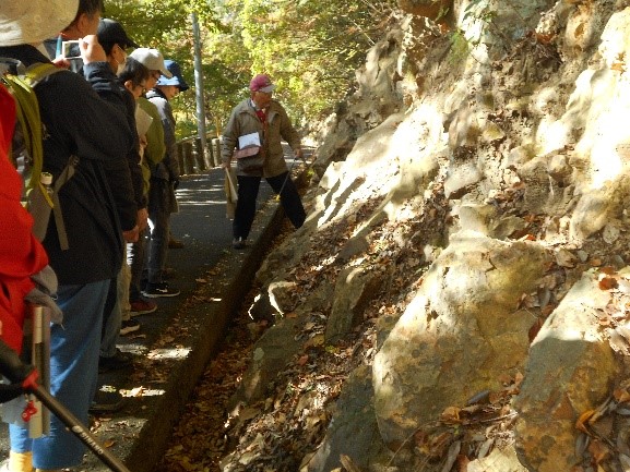 児童遊園内の歩道横の傾斜のある場所の地層について説明する講師と並んで話を聞く受講者たちを撮影した写真