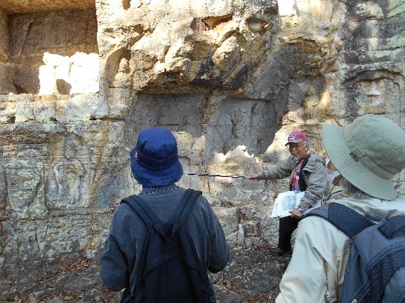 白く大きな岩谷観音磨崖仏の地層や地質について説明する講師と上を見上げる受講者たちを撮影した写真
