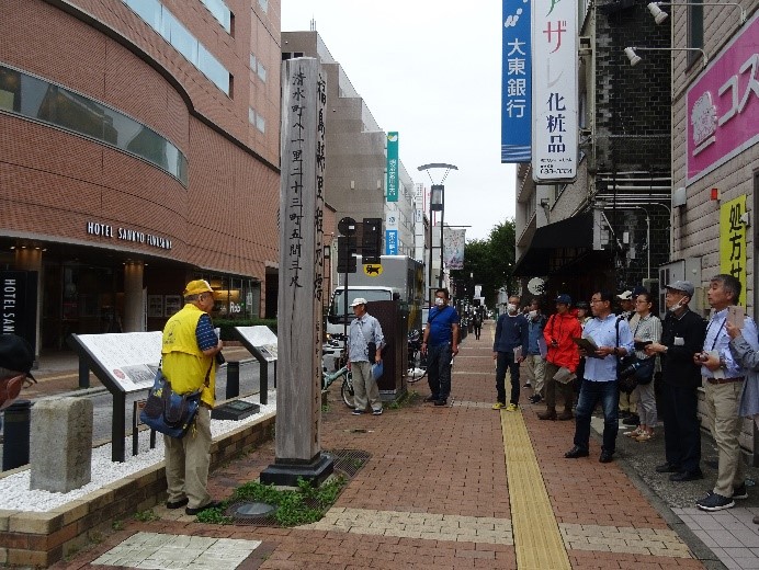 繁華街の真ん中に建てられた福島懸里程元標の角材を下から見上げる受講者たちの写真