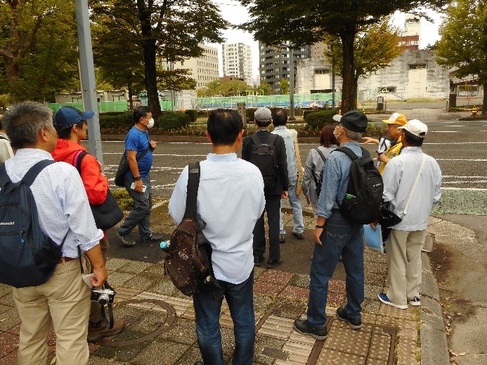 二車線道路の真ん中に置かれた福島城大手門跡の看板を道路脇に集まり講師の話を聞く受講者たち