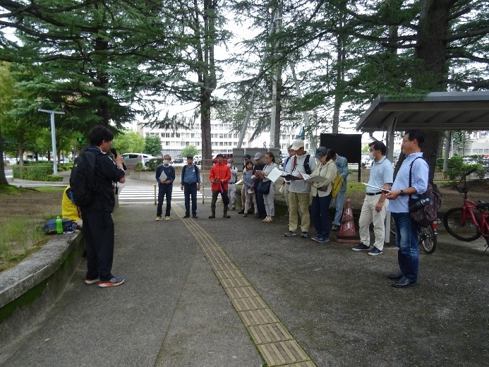 県庁の木が植えられた横のスペースに集まり締めの挨拶を聞く受講者たちを撮影した写真