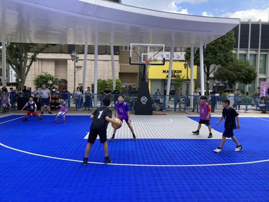 地面が青い3×3バスケットコートで黒のユニフォームを着たチームと紫色のユニフォームを着たチームが試合を行っている写真
