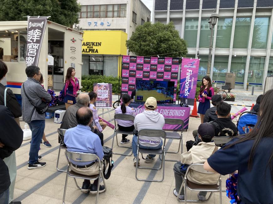 屋外の一角にモニターが設置され、設置されたパイプ椅子に参加者達が座りパブリックビューイングが行われている様子の写真