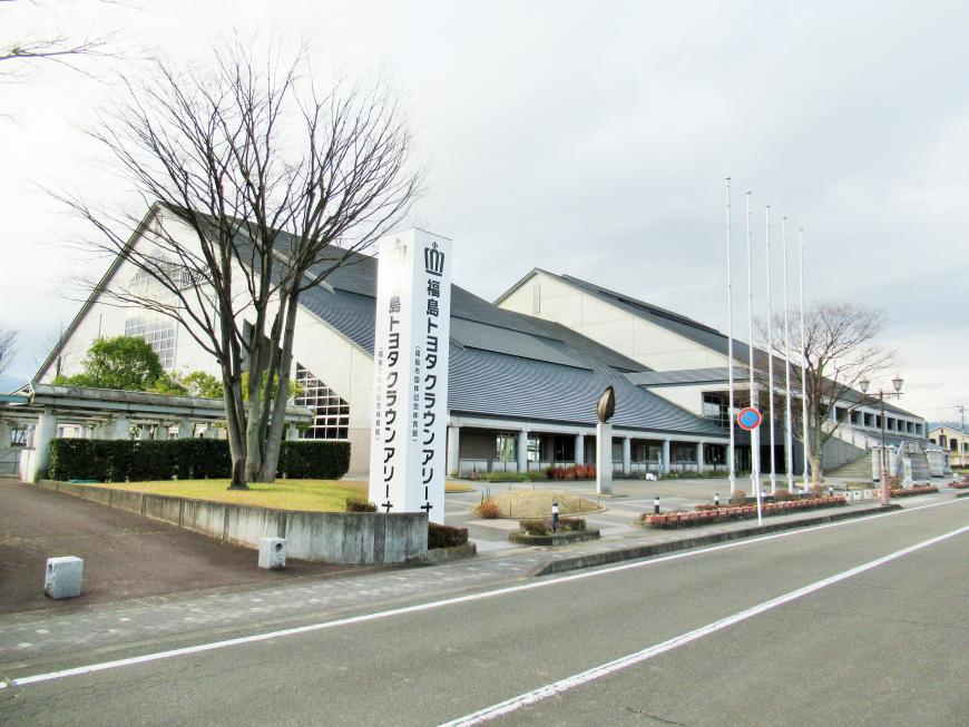 道路に面した敷地入口に「福島トヨタクラウンアリーナ」と書かれた白い柱の看板があり、奥に三角屋根の大きな建物が建っている写真
