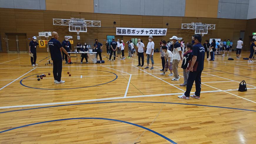 白い線に沿って参加者が横一列に並び、青い線が引かれた円の中にボールが複数入っているボッチャ交流大会が行われている様子の写真