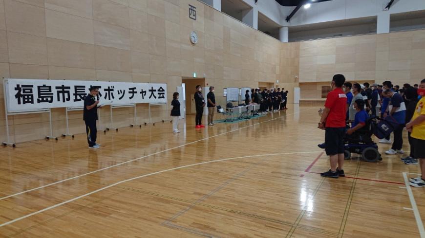 「福島市長杯ボッチャ大会」と書かれた紙がホワイトボードに貼られ、その前にマイクを持って話をしている男性、男性の向かい側に多くの参加者が整列している写真