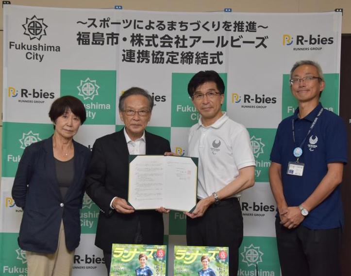 締結式でバックパネルの前に下条副社長、橋本社長、木幡市長、西坂市民・文化スポーツ部長が立ち、橋本社長と木幡市長が締結書を一緒に持ち記念撮影をしている写真