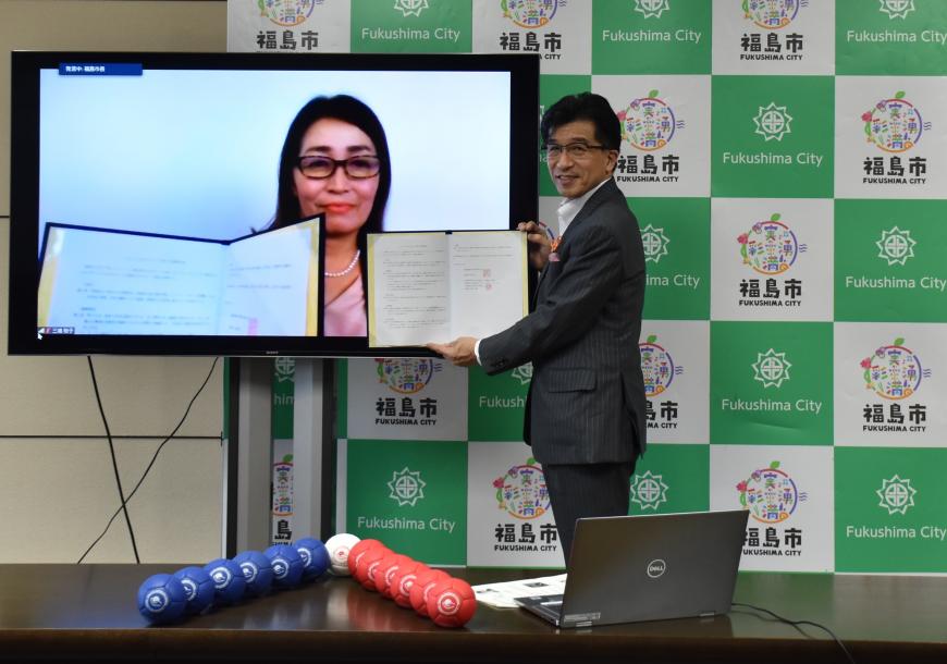 モニターに映し出された協定書を持っている一般社団法人日本ボッチャ協会三浦裕子事務局長と、モニターの横に立ち協定書を持っている木幡市長の写真