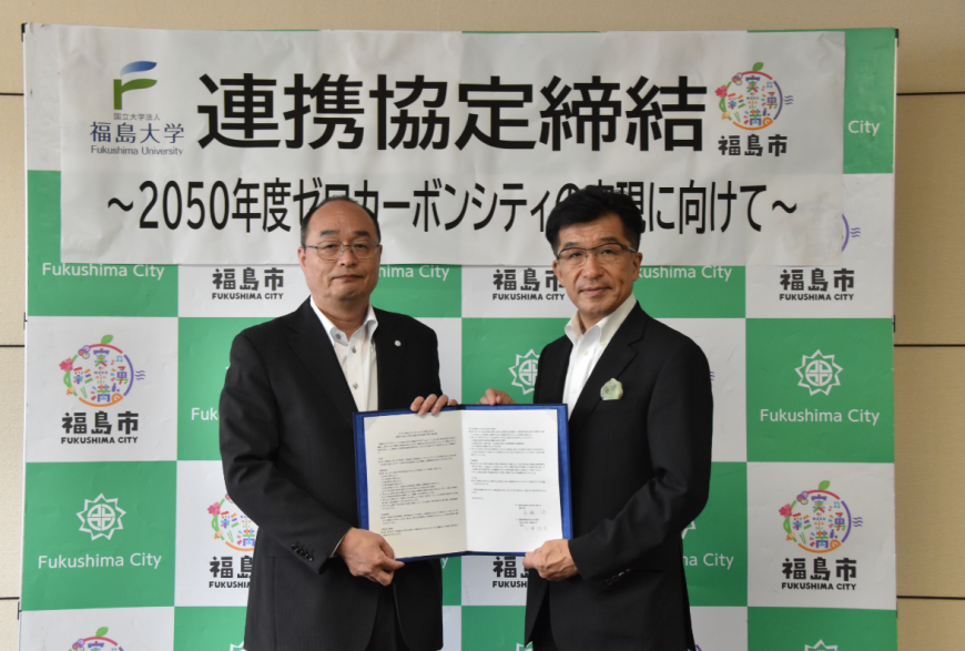 福島市のバックパネルの前に立って協定書をもってこちらに見せている福島大学代表者と木幡市長の写真