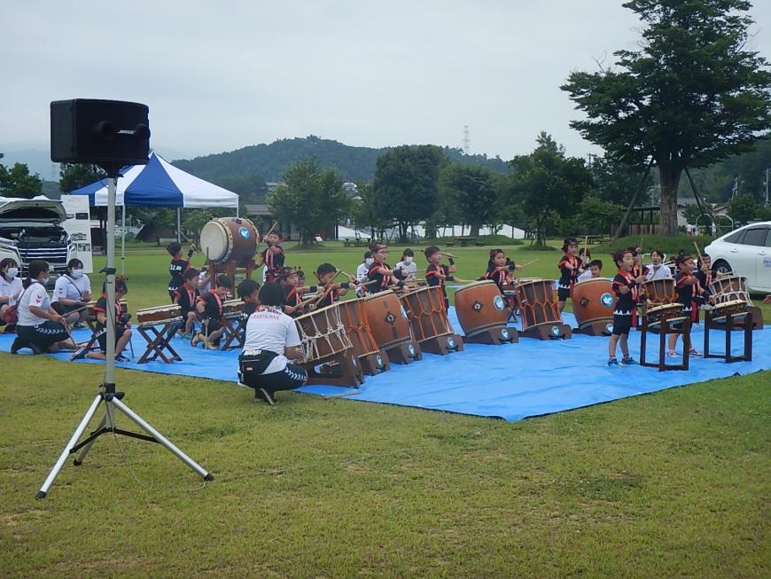 芝生に敷かれたブルーシートの上で太鼓の演奏を披露している霊山三育認定こども園の園児たちの写真