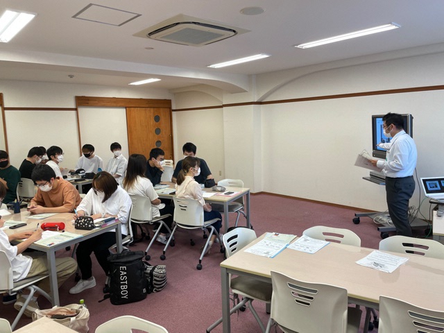 前方のモニターの横で話をしている男性とグループごとに分かれて講義を受けている参加者の写真