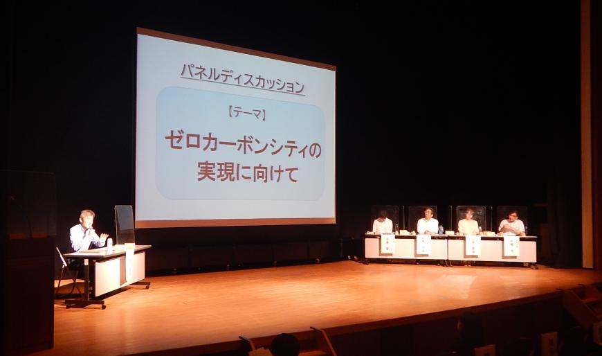 資料が投影された舞台上の大きなスクリーンの横で4名のパネリストと司会者がパネルディスカッションしている写真