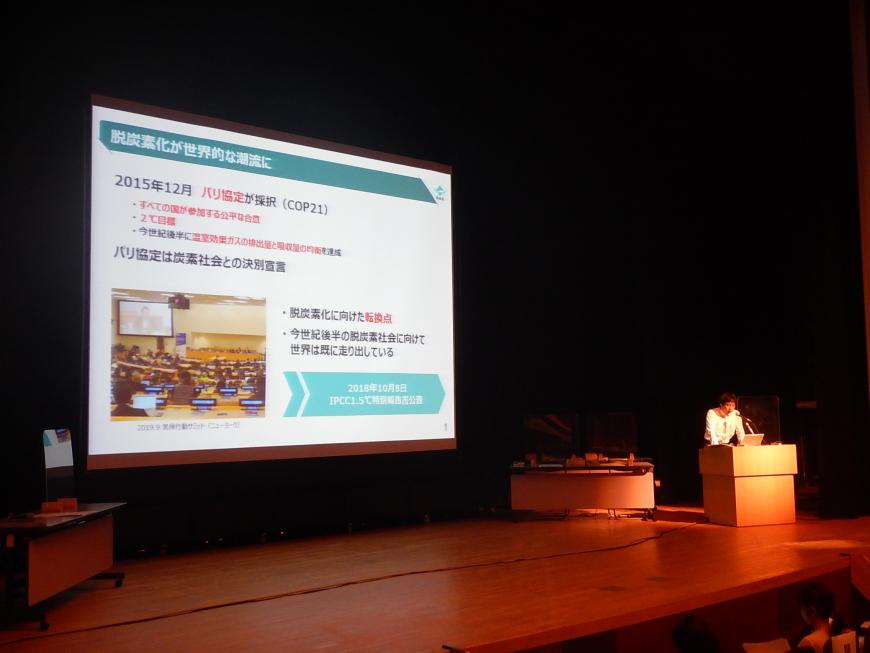資料が投影された舞台上の大きなスクリーンの横で演台に立ち話をしている加藤室長の写真
