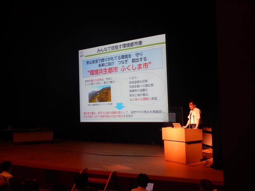 資料が投影された舞台上の大きなスクリーンの横で演台に立ち話をしている木幡市長の写真