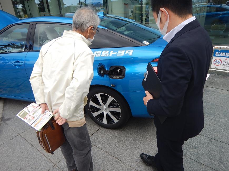 青い車体の燃料電池自動車の前でスタッフの話を聞いている男性の写真