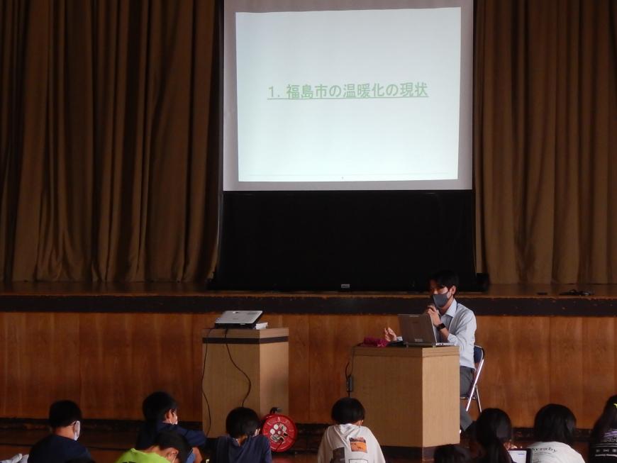 資料が投影された舞台上の大きなスクリーンの前の演台で話をしている男性と話を聞いている子供たちの写真