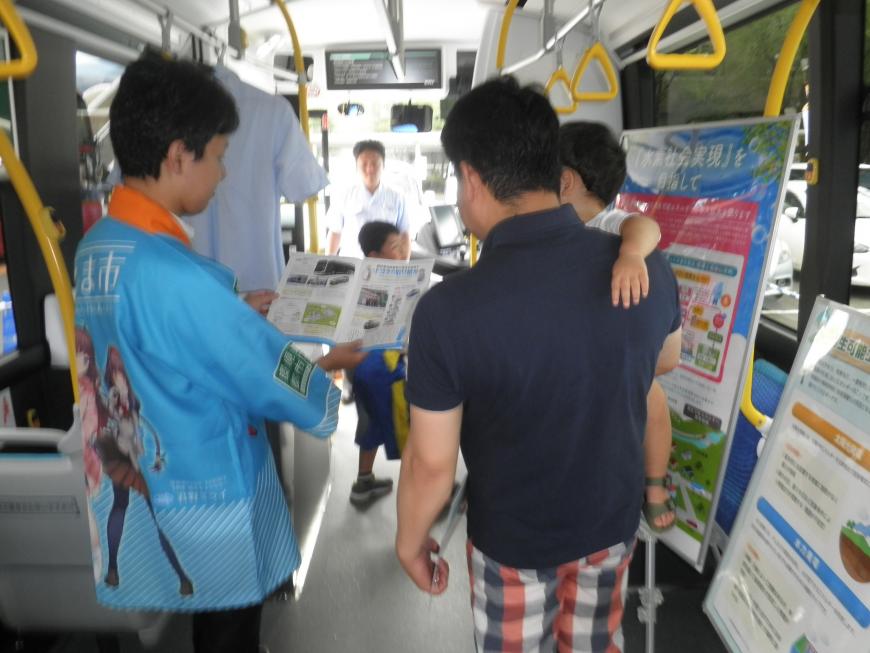 展示パネルが設置されたバスの車内でパンフレットを見せながら説明しているスタッフと話を聞いている子供を抱いた男性の写真