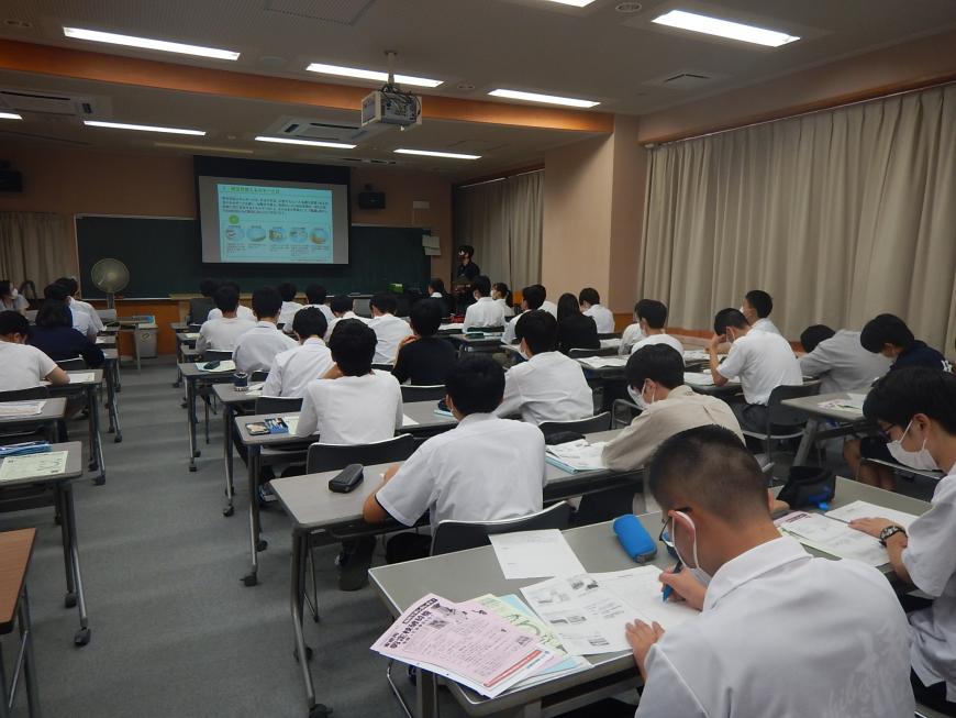 資料が投影された前方の大きなスクリーンと手元の資料を見ながら講義を受けている学生の写真