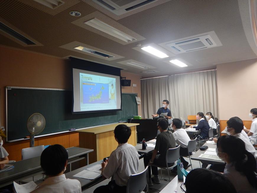 前方のスクリーンの横に立っている男性、席に着き話を聞いている学生たちの写真