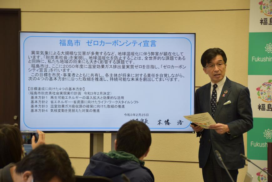 福島市ゼロカーボンシティ宣言の内容が表示されたモニターの横に立って話をしている市長の写真