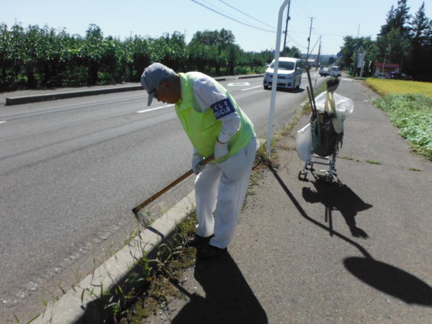 帽子を被った作業着姿の男性が歩道から道路脇の雑草を鍬を使って取っている様子の写真