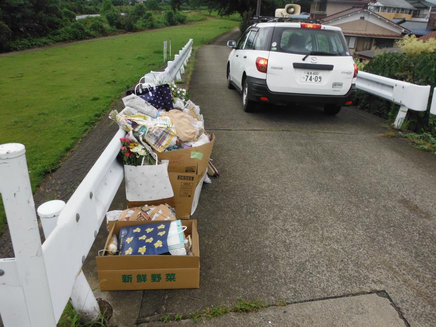 道路沿いのガードレールの前に段ボールいっぱいにお盆の供物が入っている状態の写真
