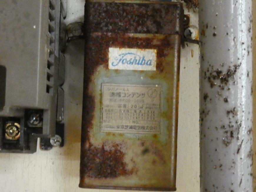 配管の横の壁に取り付けられている一部容器がさびている長方形の形をしたコンデンサの写真