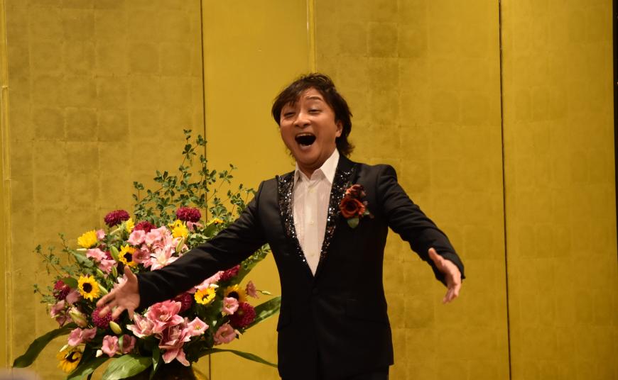 金屏風の前に立ち、世界的オペラ歌手の樋口達哉さんが両手を広げて歌を歌っている写真