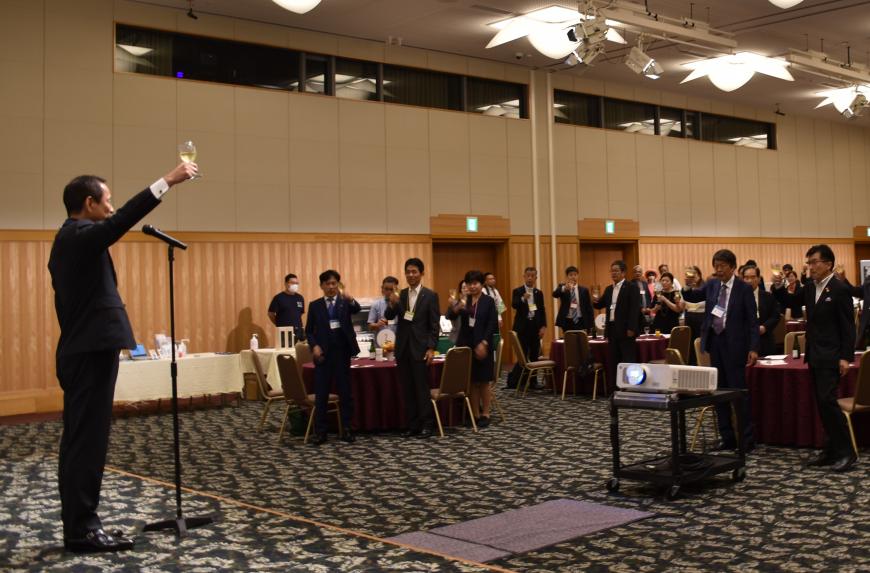 グラスを持った右手を上に上げ、参加者達に向かって乾杯の発声をしている横山淳委員の写真