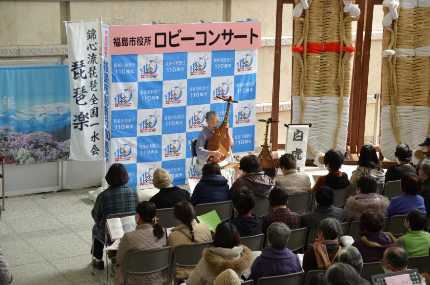 「福島市役所ロビーコンサート」と書かれたバックパネルの前に座った薄紫色と白のグラデーションの着物を着た女性が琵琶を演奏している写真
