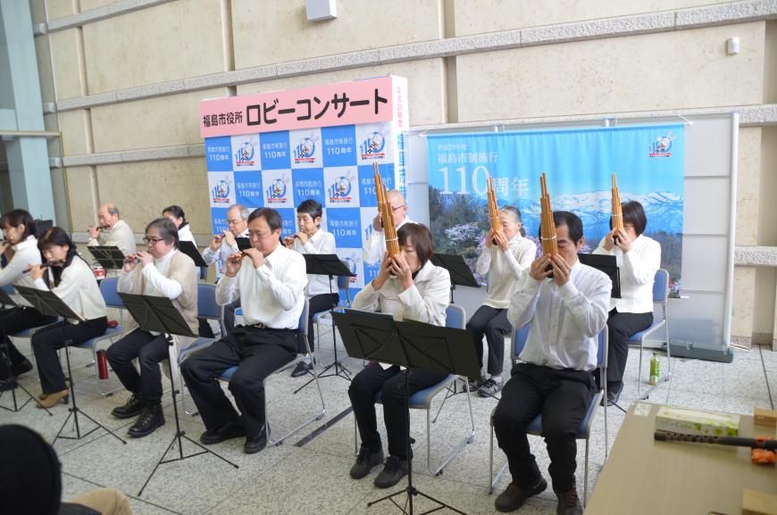 「福島市役所ロビーコンサート」と書かれたバックパネルの前に2列に並んで座った男女が様々な管楽器で雅楽の演奏をしている様子を右側から写した写真