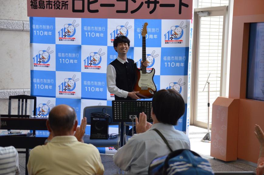 「福島市役所ロビーコンサート」と書かれたバックパネルの前でギターを両手で持った味曽野さんが笑顔で客席を向いている写真