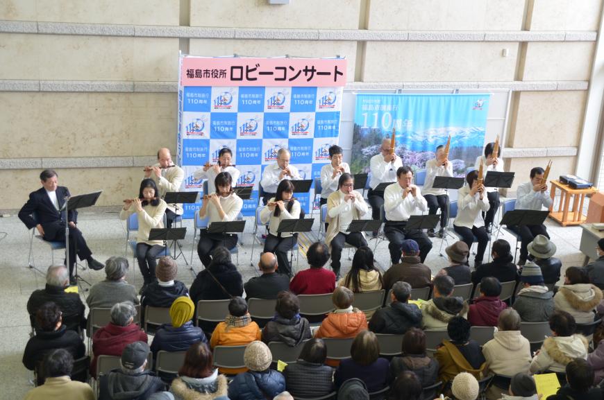 たくさんの来場者が客席に座る中、「福島市役所ロビーコンサート」と書かれたバックパネルの前に2列に並んで座った男女が様々な管楽器で雅楽の演奏をしている写真