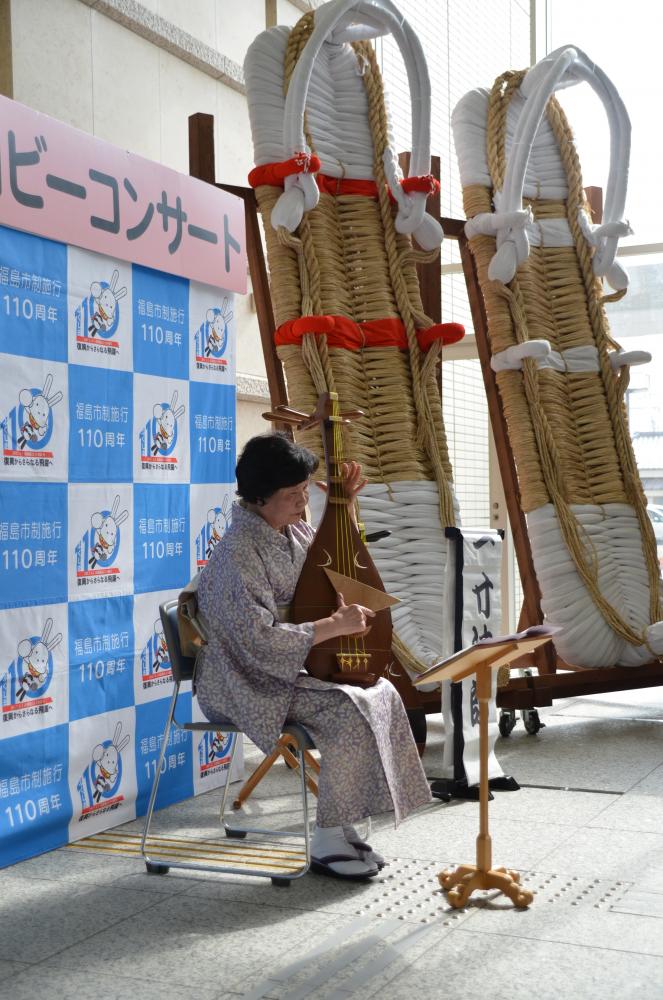 バックパネルの前に座った薄紫色の着物を着た女性が琵琶を演奏している写真