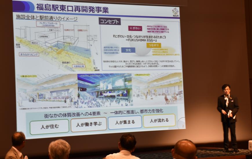ステージ上のスクリーンに映し出された資料の横に立ち、木幡市長が挨拶をしている写真