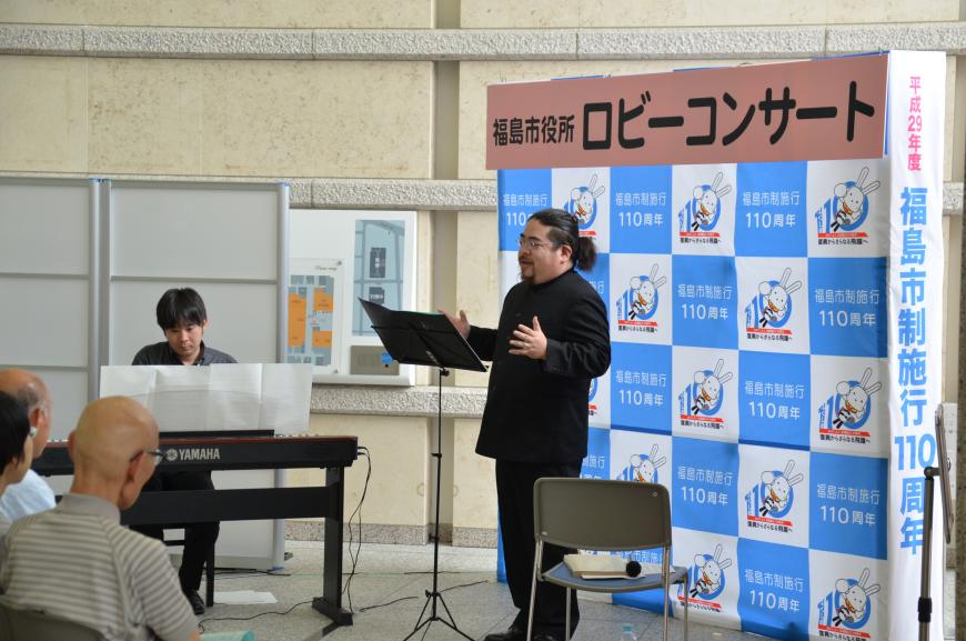 左側に置かれた電子ピアノを弾く男性と、「福島市役所ロビーコンサート」と書かれたバックパネルの前で両手を広げて歌唱している黒い衣装の男性の写真