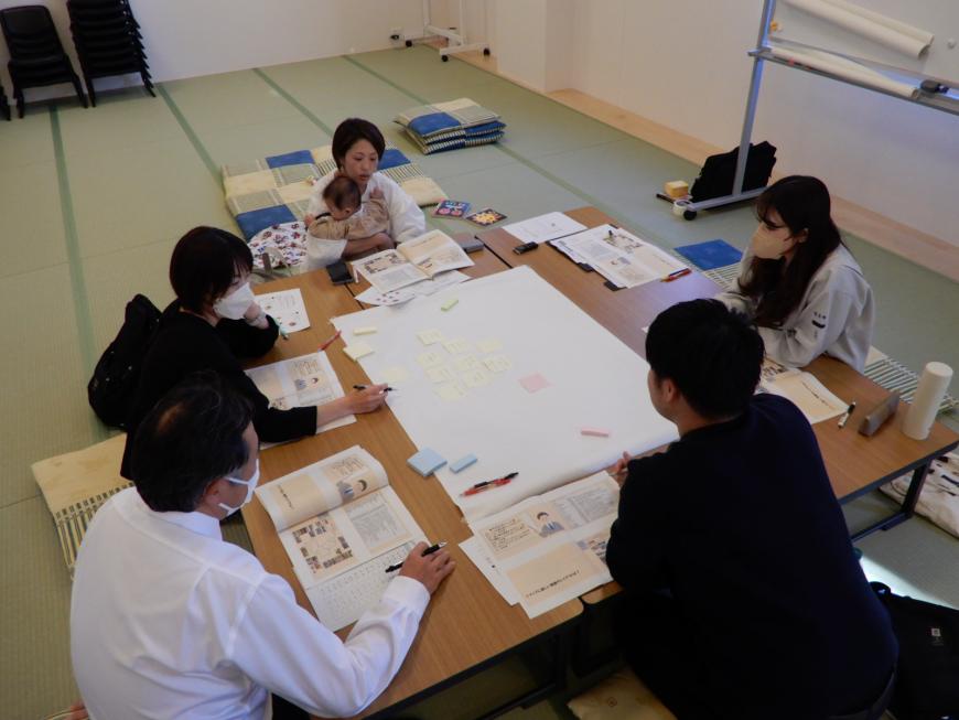 8名の関係者の方々が座りながら四角の大きめのテーブルの中央に付箋を貼ったりして話し合いをしている写真