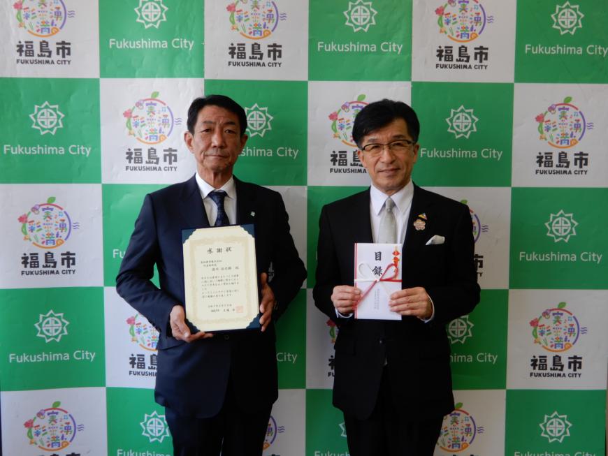 バックボードの前で市長が目録を持ち、恵和興業株式会社の男性が感謝状を持ち2人で記念撮影をしている写真