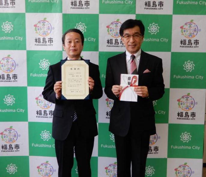 バックボードの前で市長が目録を持ち、ジーエルサイエンス株式会社の男性が感謝状を持ち2人で記念撮影をしている写真