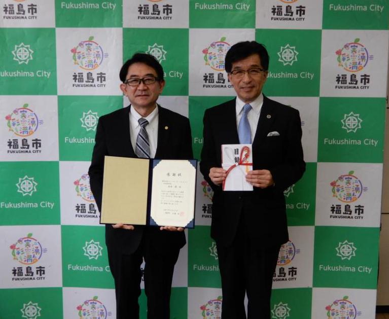バックボードの前で市長が目録を持ち、ファーマーズ・フォレストの男性が感謝状を持ち2人で記念撮影をしている写真