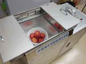 箱形で銀色をした非破壊放射能測定装置の中にザルに入ったリンゴ6個が入れられている写真