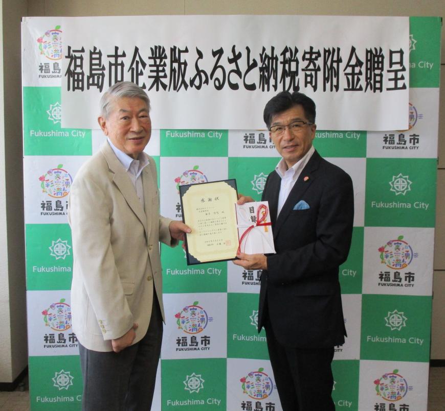 バックボードの前で市長と株式会社コミューンの男性が感謝状と目録を2人で持ちながら記念撮影をしている写真