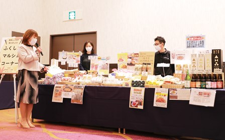 沢山の商品が陳列された台の横にマイクを持った女性が立ち話をしている様子の写真