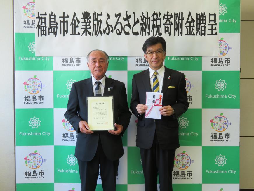 バックボードの前で市長が目録を持ち、株式会社パスクの男性が感謝状を持ちながら2人並んで記念撮影をしている写真