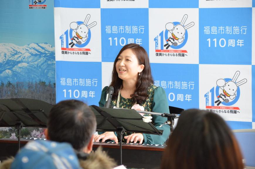 福島市制施行110周年のバックパネルの前に設置された電子ピアノの演奏席に座った星さんが笑顔で演奏をしている写真